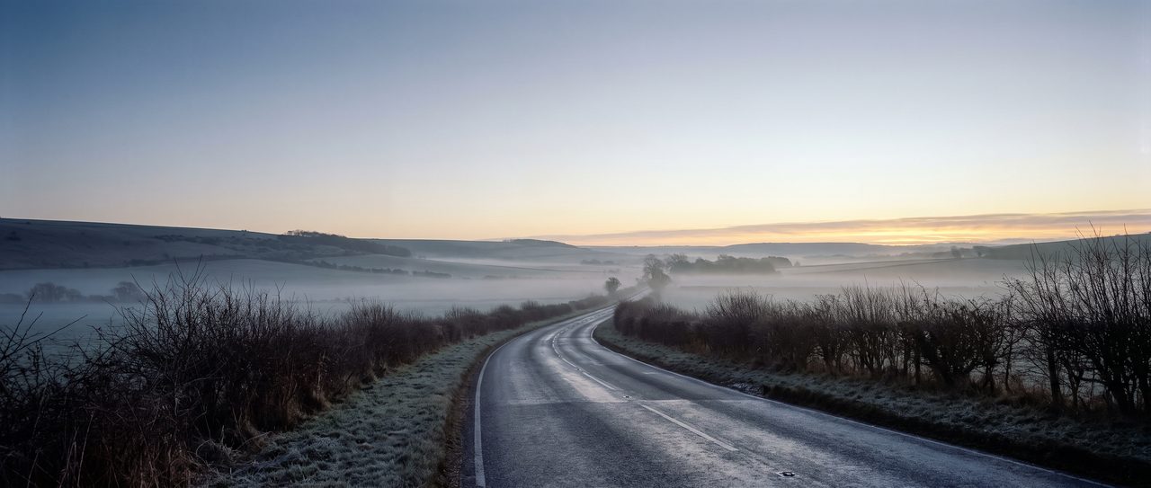 An empty country road at first light, mist lying across the fields